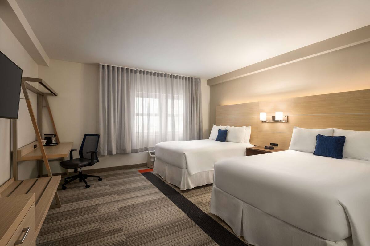 A hotel room with two beds, a desk, a chair, a TV, and a window with curtains.