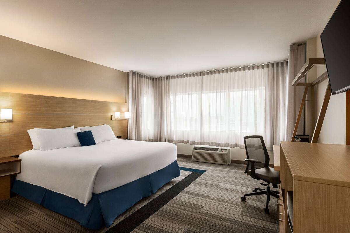 A hotel room with a large bed, a desk, a chair, a TV, and a window with curtains.
