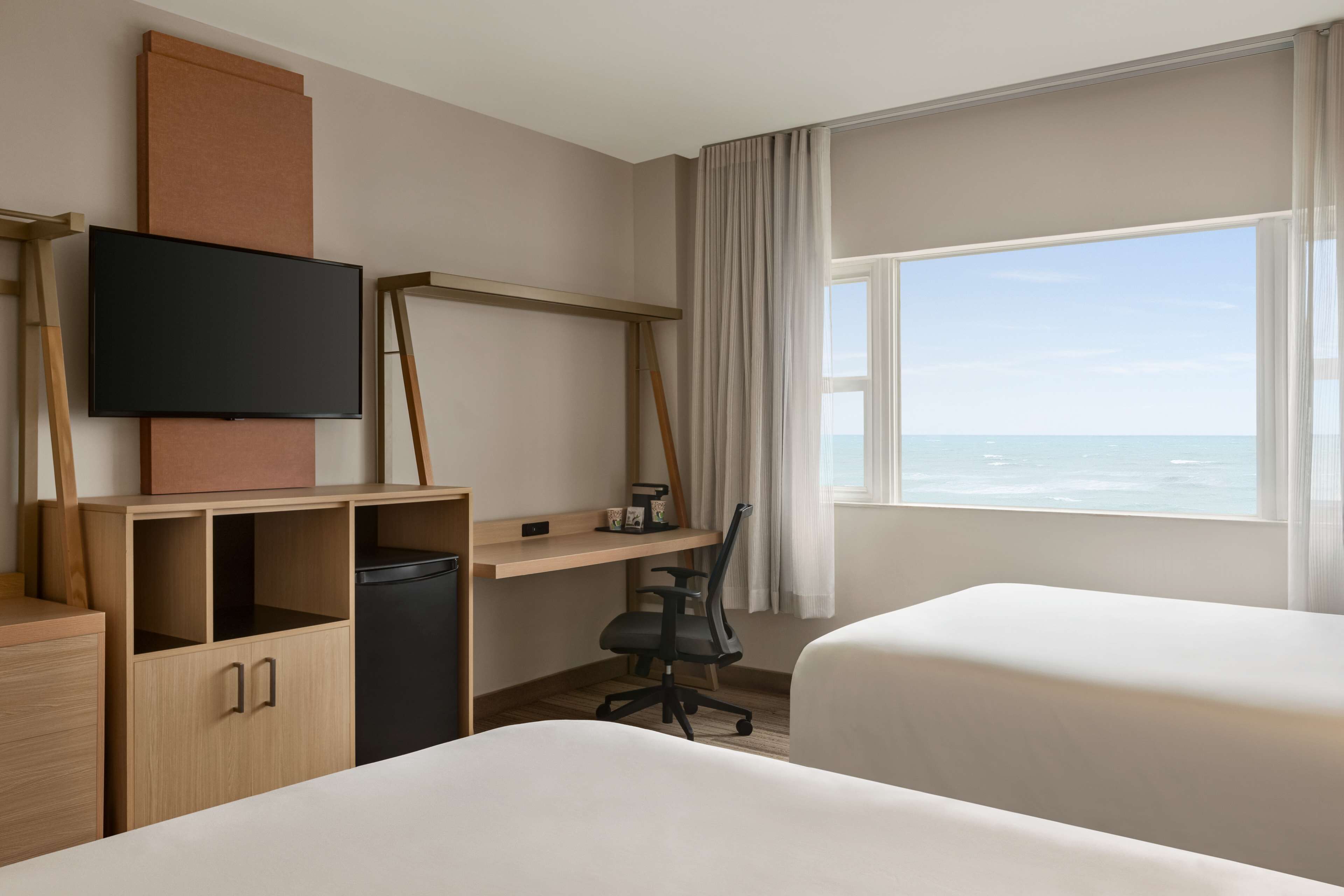 Een hotelkamer met een bed, een bureau met stoel, een televisie en een raam met uitzicht op de oceaan.