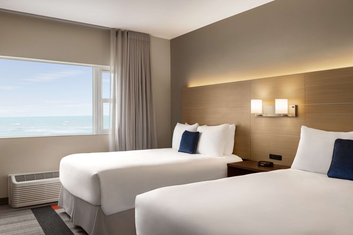 Una habitación de hotel con dos camas, un ventanal con vista al océano, un aplique en la pared y un calentador.