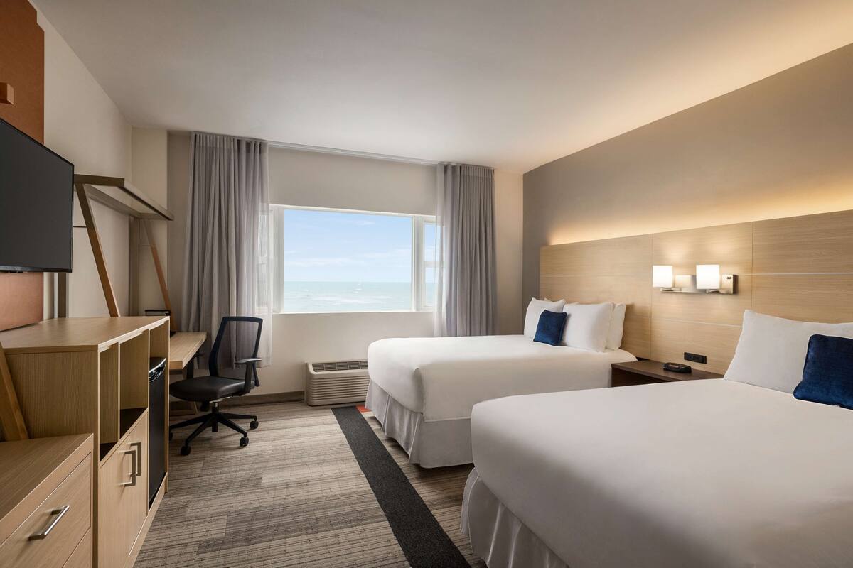 A hotel room with two beds, a desk, a chair, a television, and a large window with curtains.