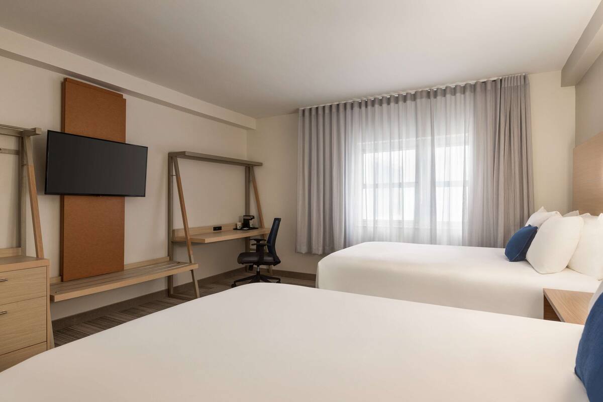 A hotel room with two beds, a desk with a chair, a flat-screen TV mounted on a wooden panel, and a large window with sheer curtains.