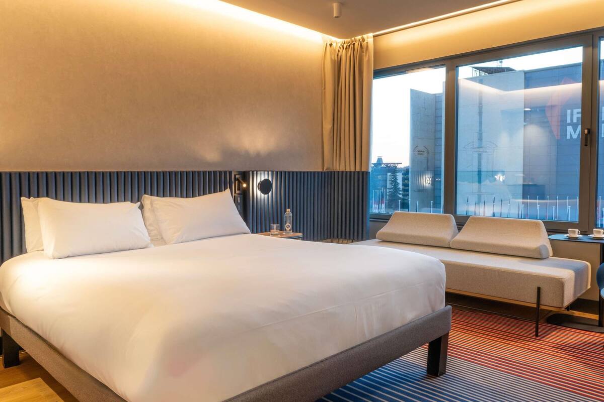 A modern hotel room with a large bed, a sofa, a small table, and a large window offering a city view.