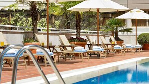 Un área junto a la piscina con sillones de descanso y sombrillas, una piscina y un edificio al fondo.