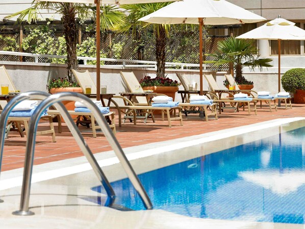 Ein Poolbereich mit Liegestühlen und Sonnenschirmen, ein Schwimmbecken und ein Gebäude im Hintergrund.