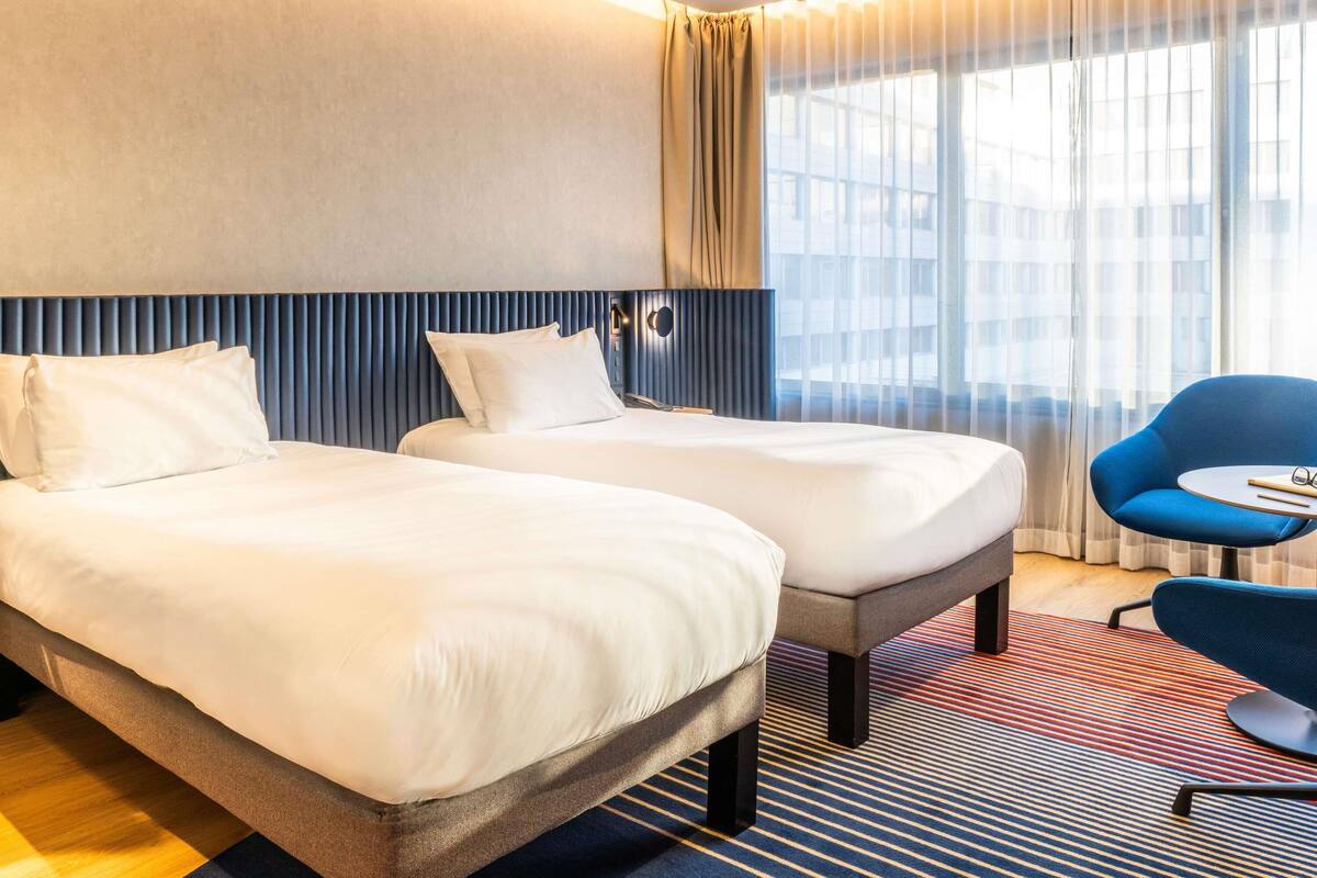 Et hotelværelse med to senge, en blå stol og et stort vindue med gardiner.