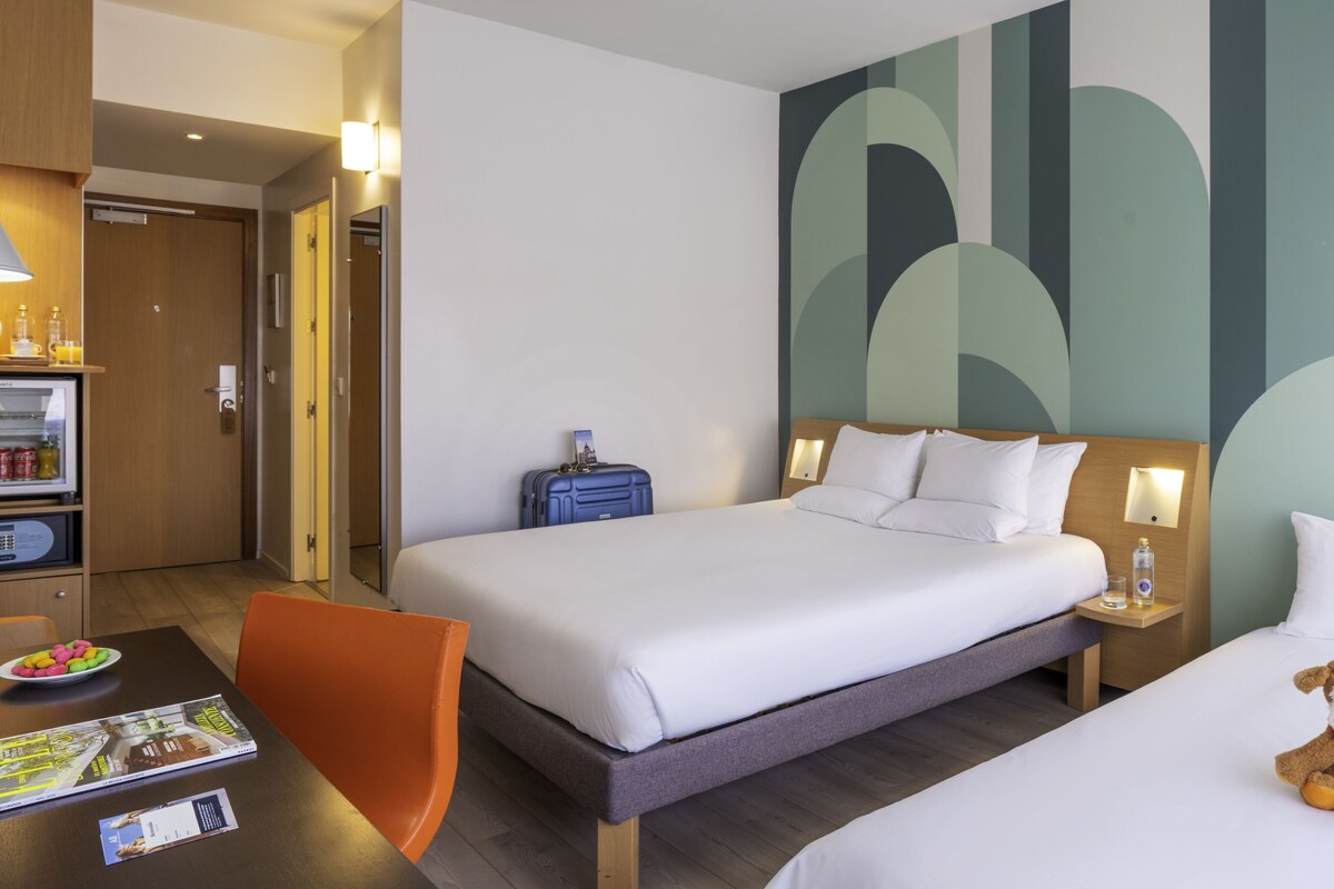 A hotel room with a bed, a desk with a chair, a small table with a plate of snacks, a suitcase, and a teddy bear on the bed.
