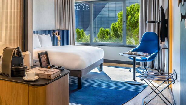 Habitación de hotel con cama, escritorio con cafetera, una silla y vista a árboles a través de ventanas grandes.