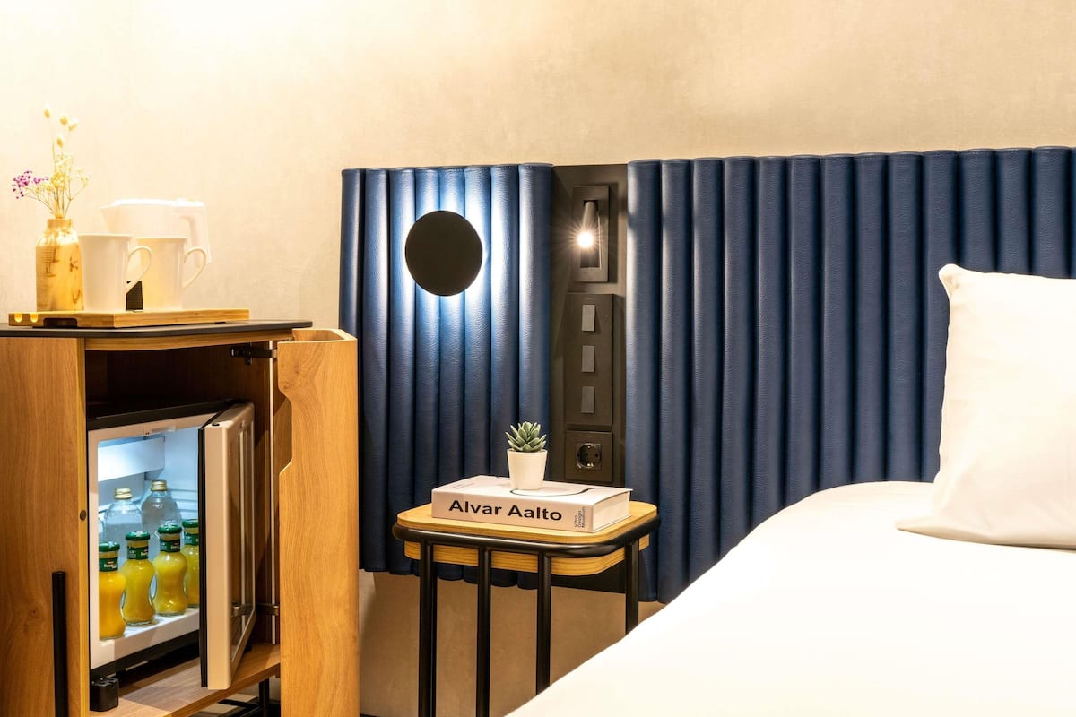 Une chambre d’hôtel avec un mini-bar, une petite table avec un livre et une plante, et un lit avec un oreiller.