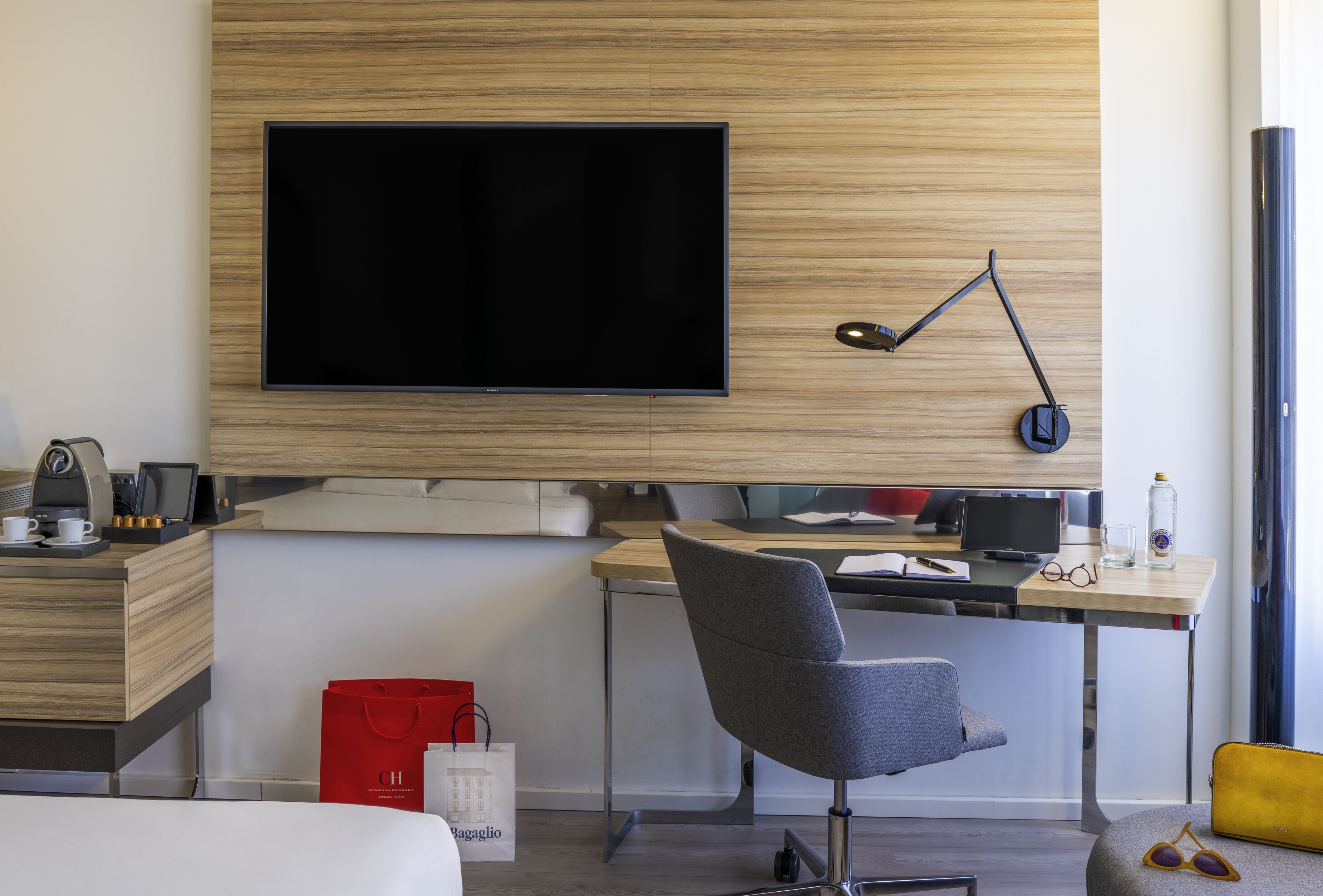 Una habitación de hotel moderna con una televisión de pantalla plana grande montada en la pared, un escritorio con una computadora, una silla y una mesita de noche con una lámpara.