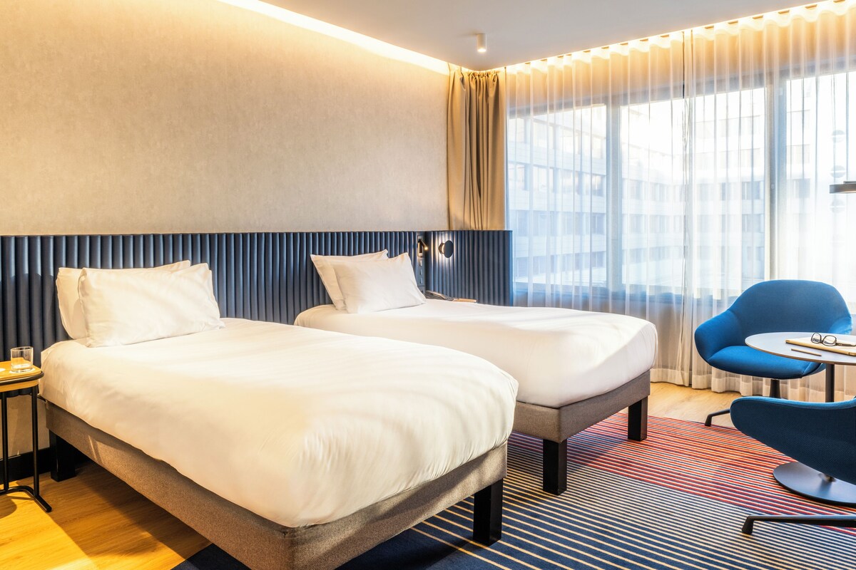 A hotel room with two beds, a blue chair, a small table, and a large window with curtains.
