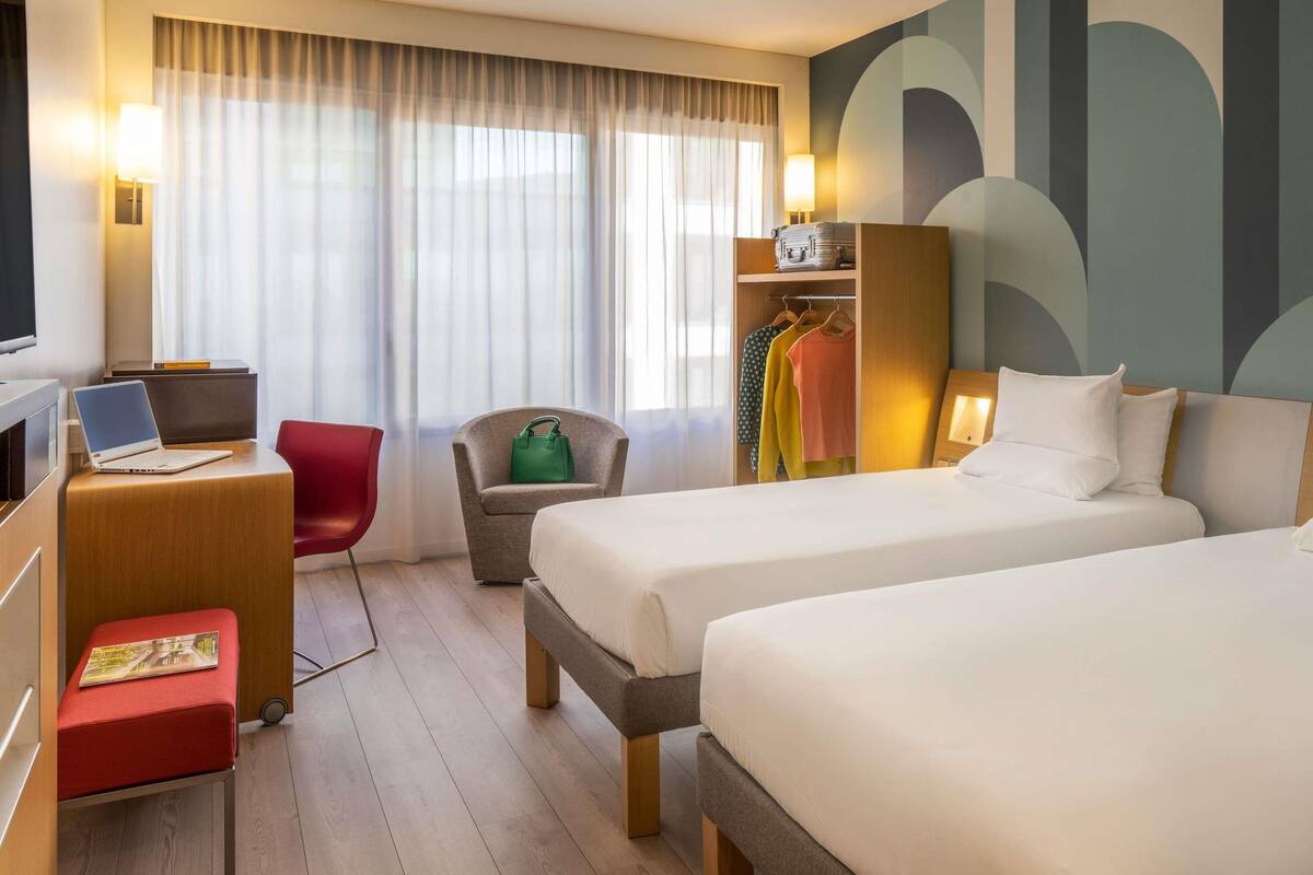 Et hotelværelse med to senge, et skrivebord med en bærbar computer, en rød stol, en grå lænestol, et klædeskab og et stort abstrakt vægdesign.