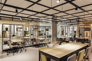 A modern conference room with multiple tables and chairs, featuring a grid ceiling with exposed beams and pendant lighting.