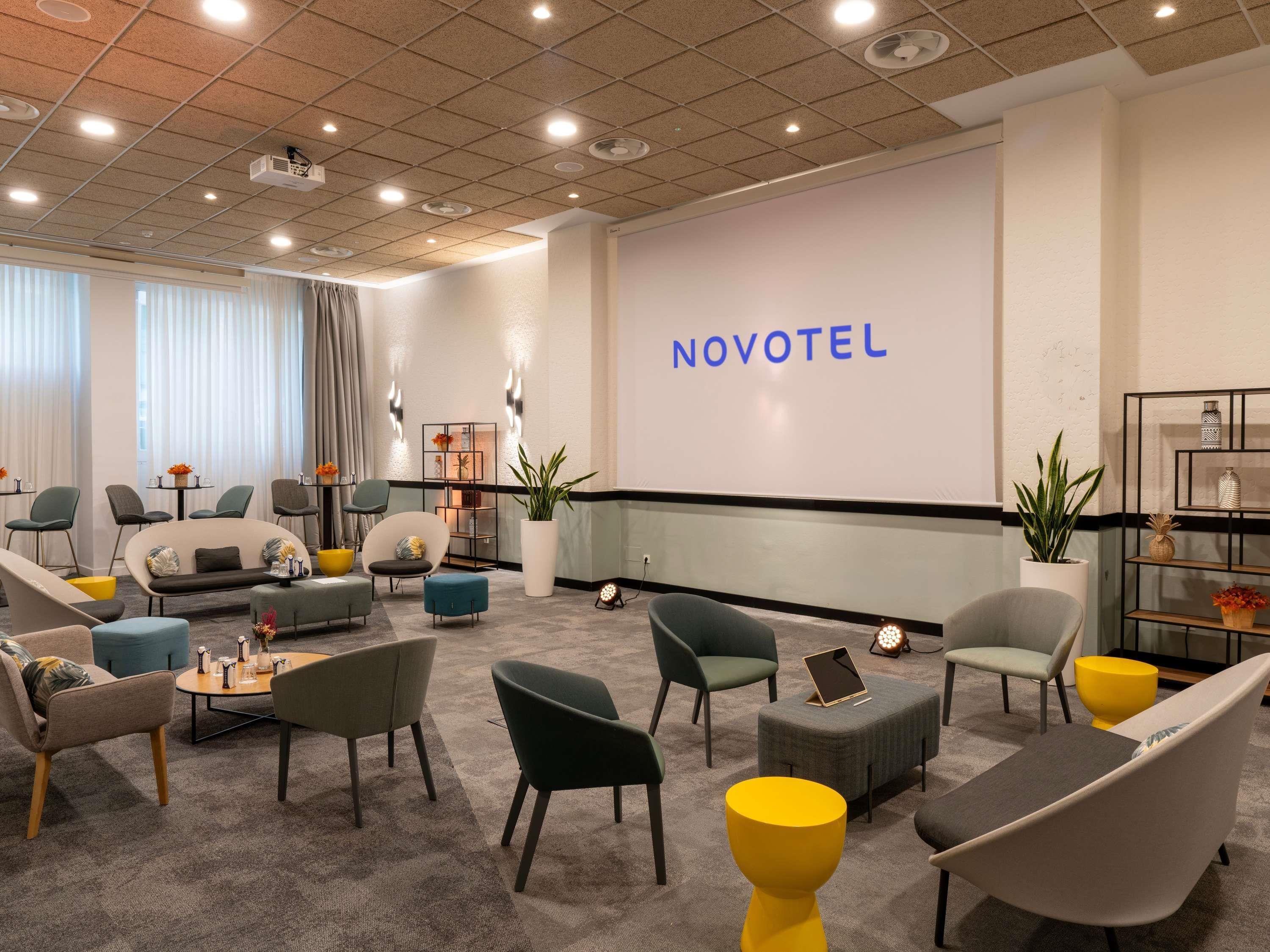 A modern hotel lobby with seating, a large screen displaying 'NOVOTEL,' and a reception desk.