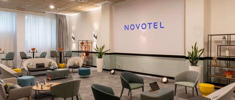 A modern hotel lobby with seating, a large screen displaying 'NOVOTEL,' and a reception desk.