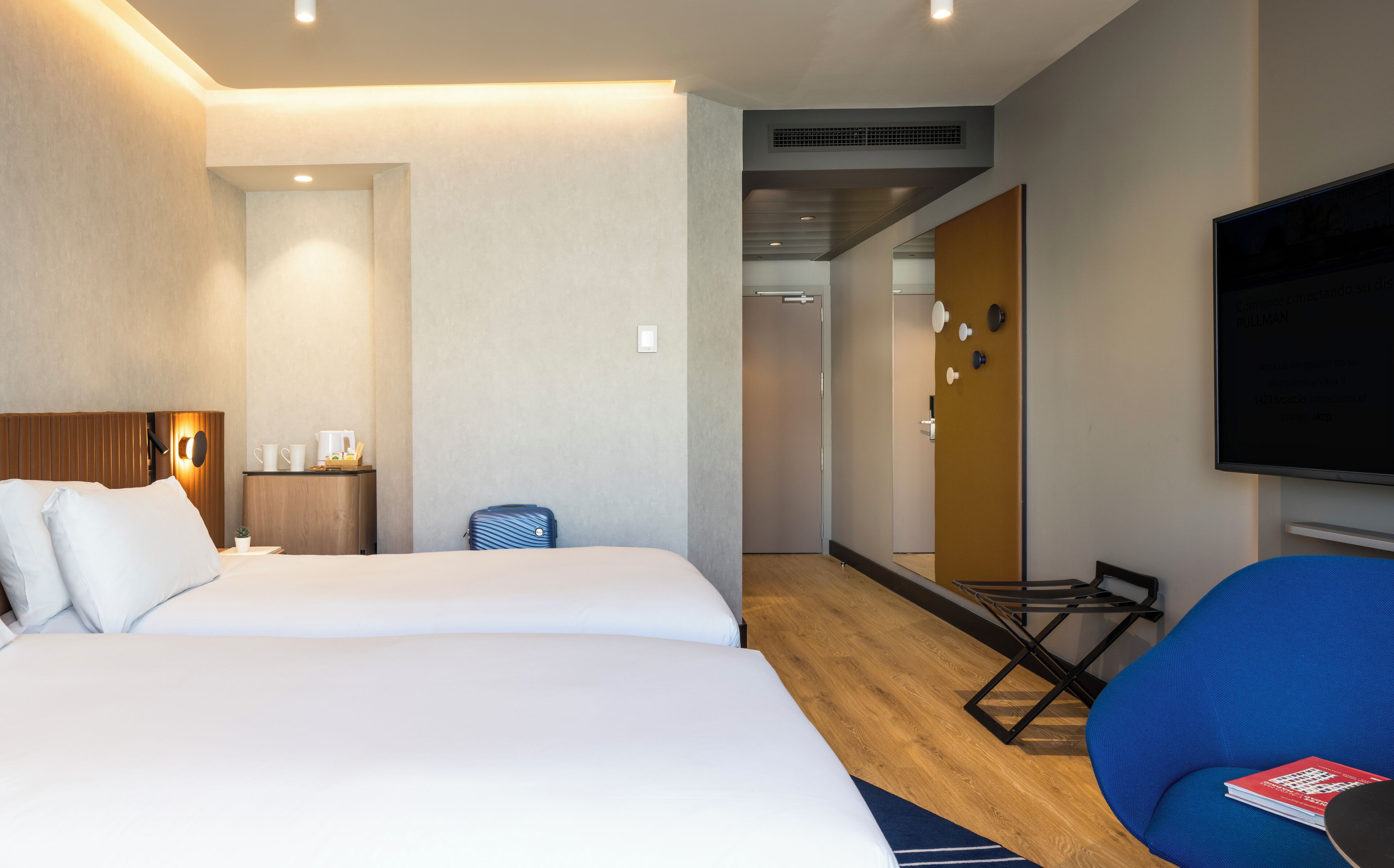 Una habitación de hotel con una cama grande, un escritorio con cafetera, una maleta azul, un televisor y una silla azul.