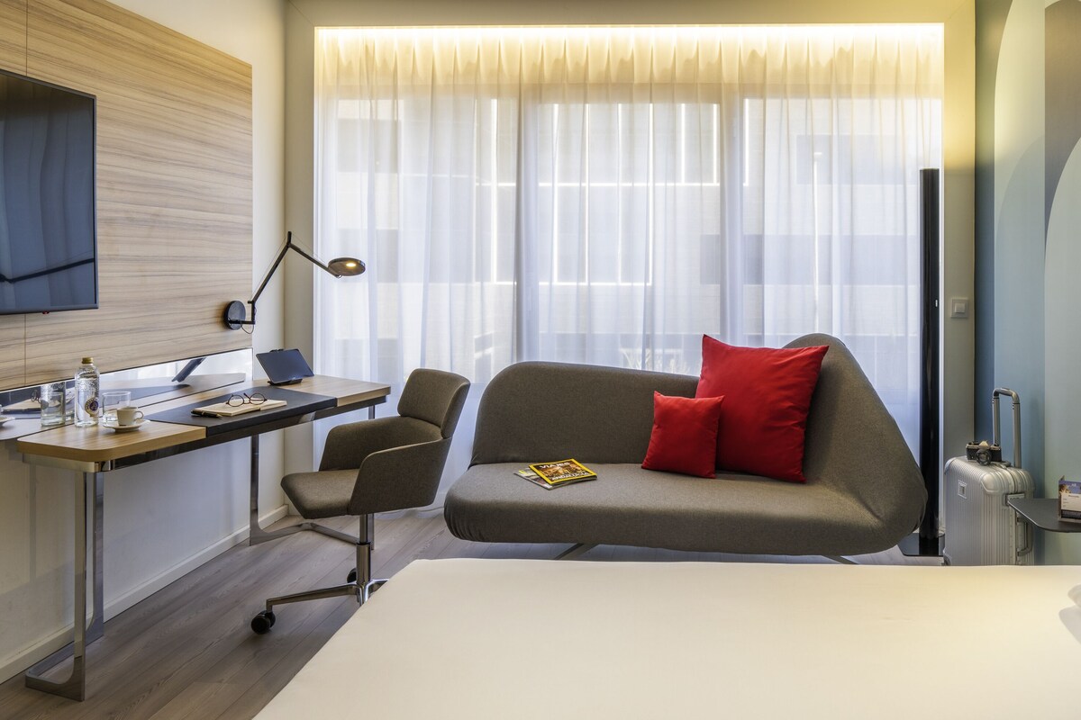 A modern hotel room with a bed, a desk with a computer, a chair, a sofa with a red pillow, a TV mounted on the wall, and a window with sheer curtains.