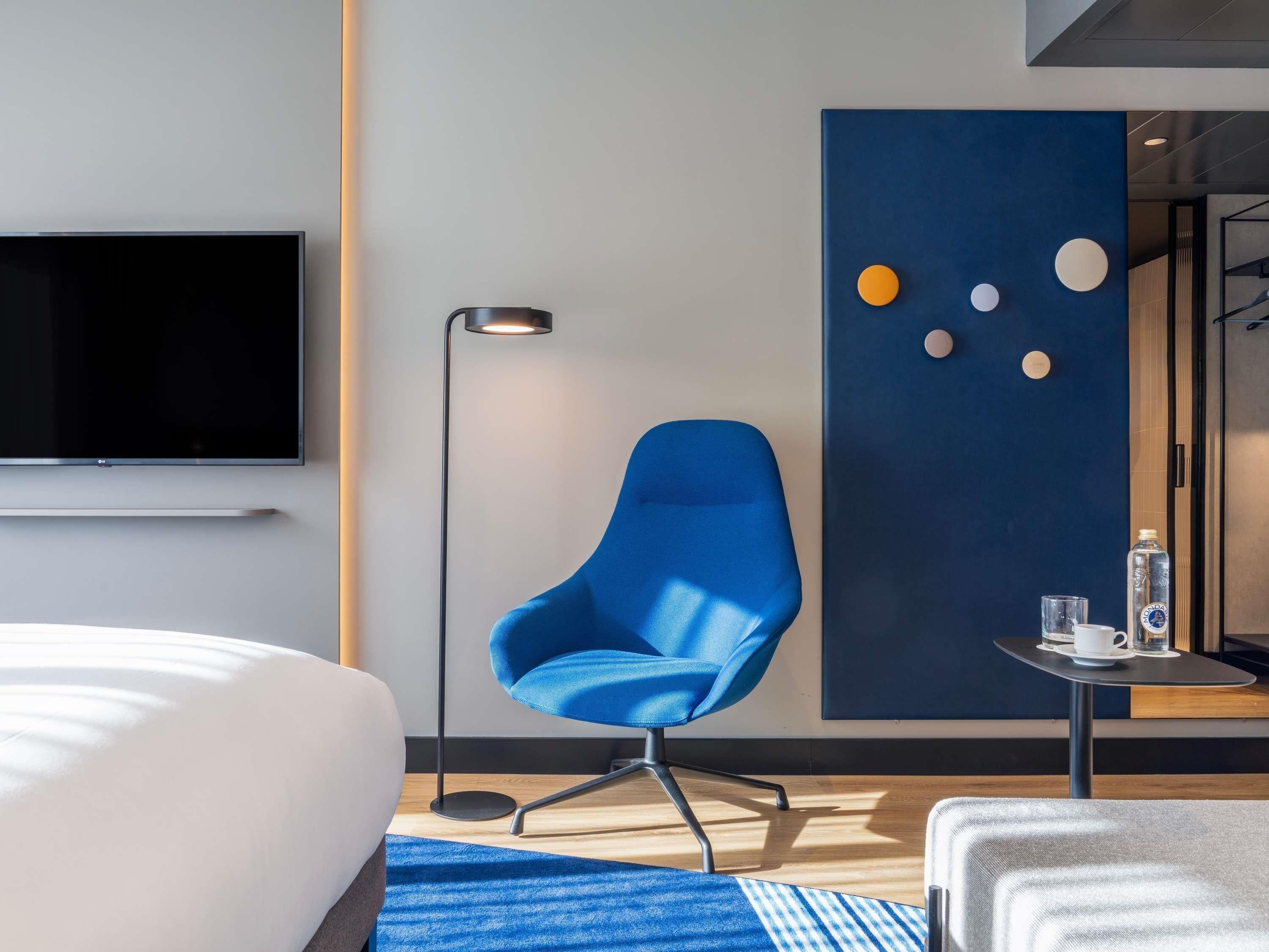 Quarto de hotel moderno com uma cama grande, uma poltrona azul, um abajur, uma pequena mesa com uma garrafa de água e uma TV fixada na parede.