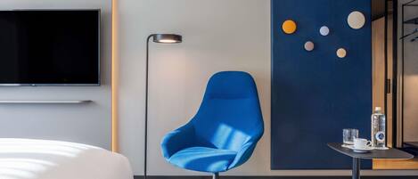 Quarto de hotel moderno com uma cama grande, uma poltrona azul, um abajur, uma pequena mesa com uma garrafa de água e uma TV fixada na parede.
