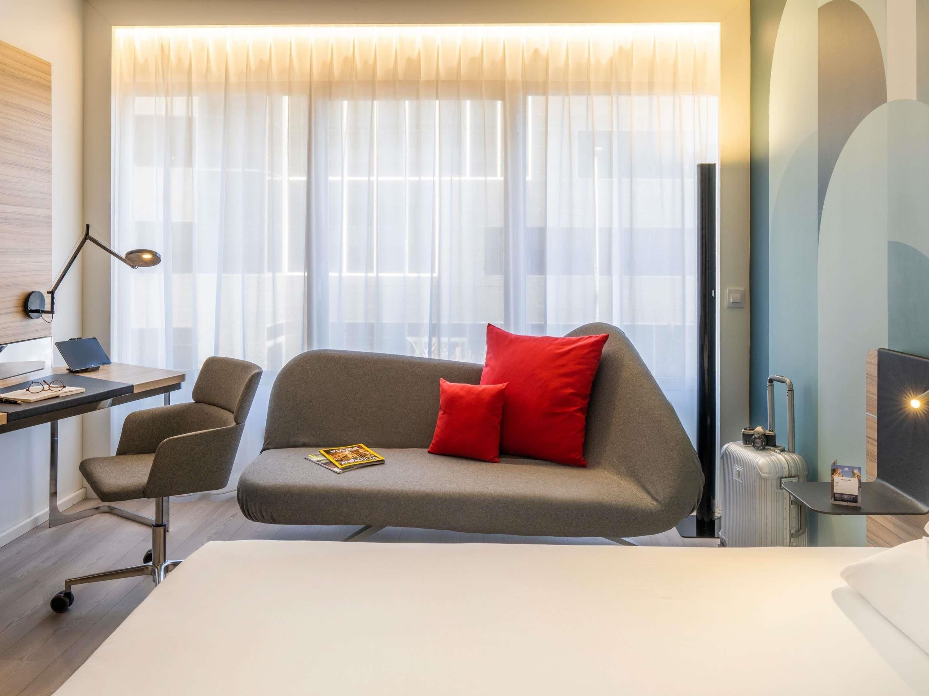 A hotel room with a bed, a sofa with red cushions, a desk with a laptop, a chair, and a suitcase.