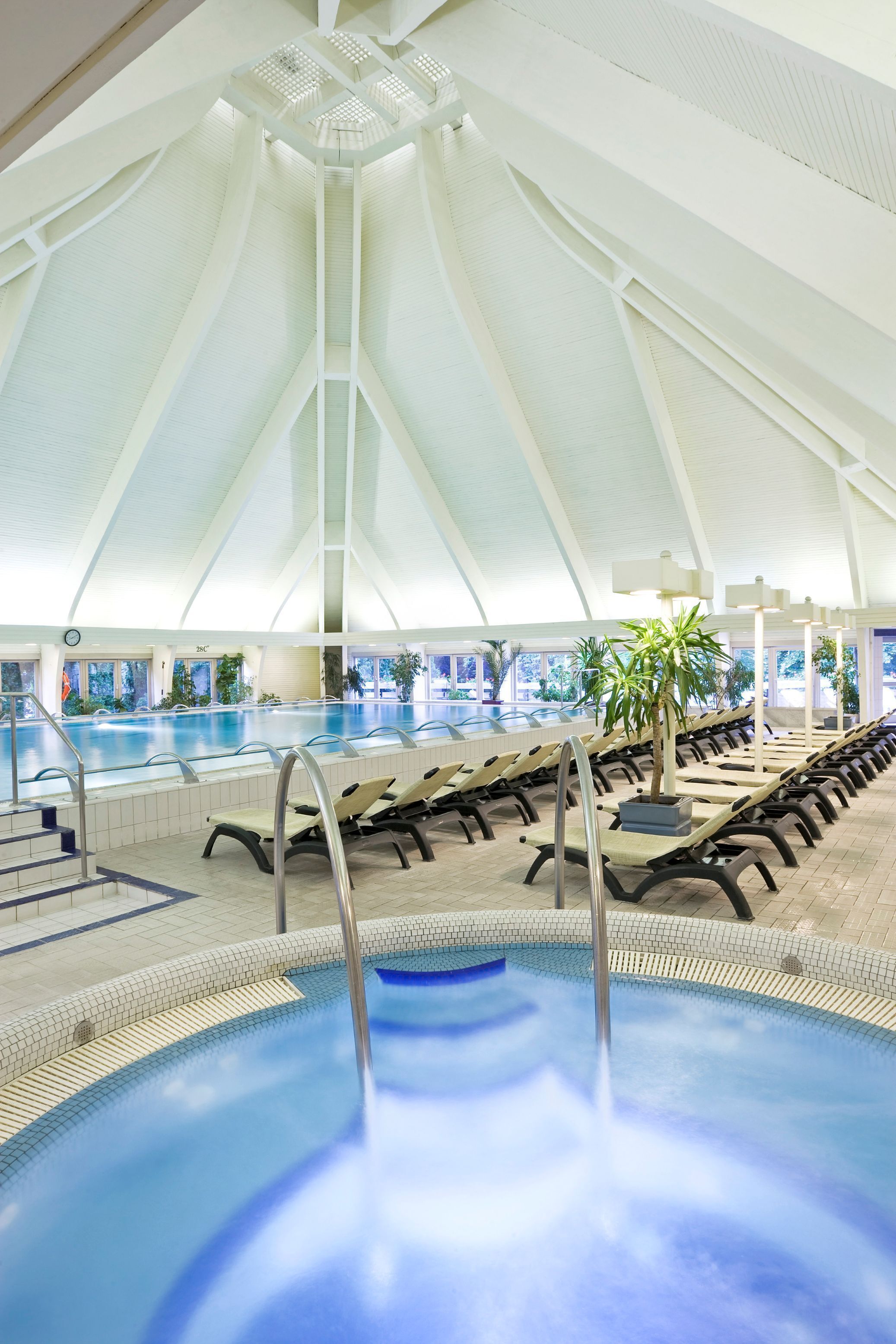 A large indoor swimming pool with a high, white, tent-like ceiling, multiple lounge chairs, and a few potted plants.