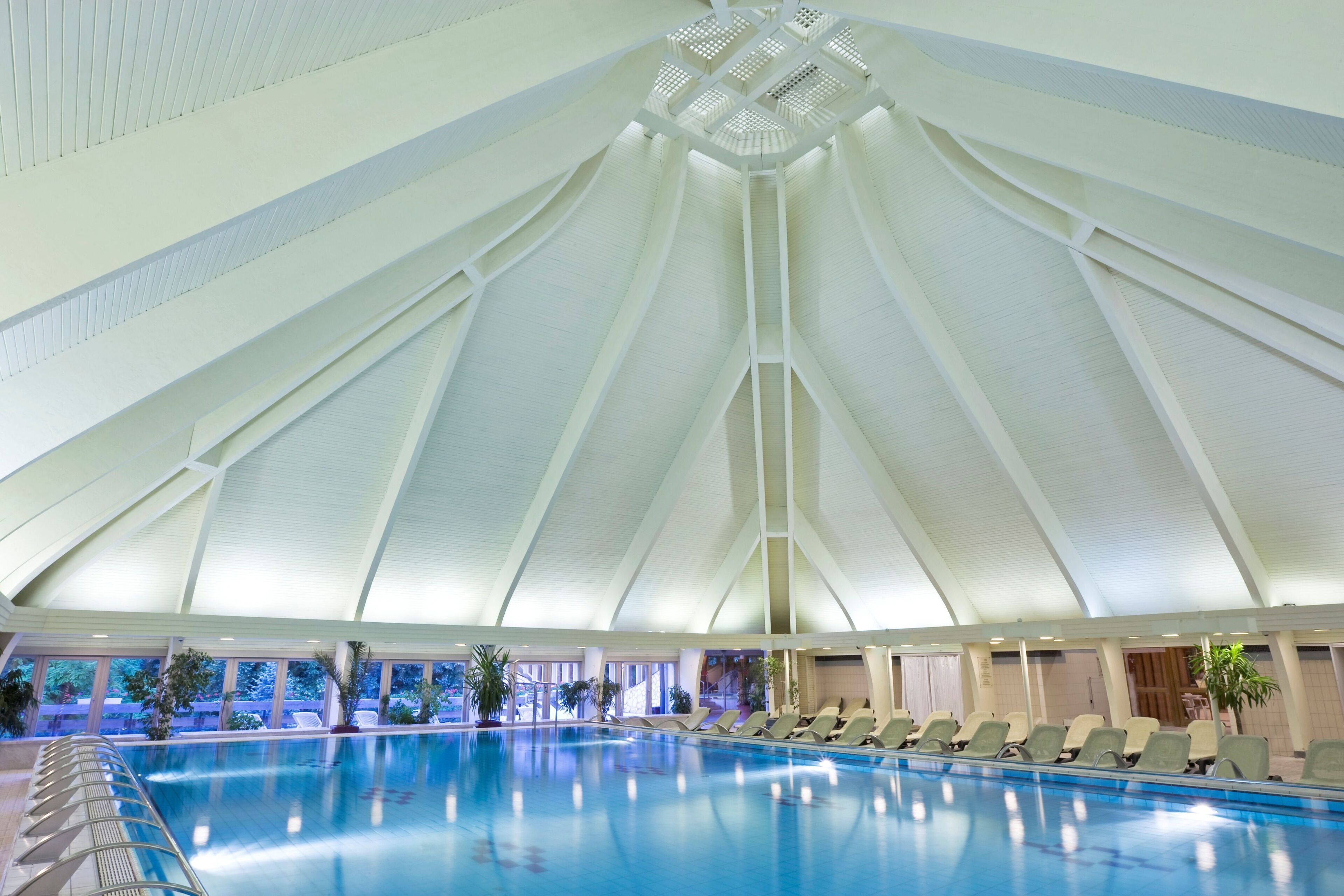 Una piscina interna con un soffitto geometrico alto e bianco e poltrone relax a bordo vasca.