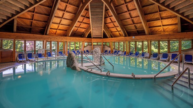 Una piscina interior con techo de estructura de madera, agua azul cristalina y sillones de descanso azules en el borde.