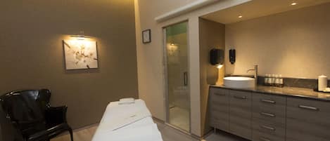 Una habitación de hotel moderna con una cama grande, una mesita de noche, una silla, un baño con lavabo y espejo, y un reloj de pared.