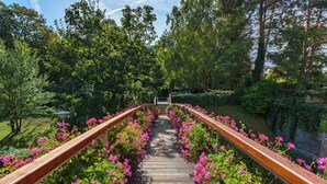 Un puente de madera con barandilla de metal, rodeado de abundante follaje y flores rosadas.