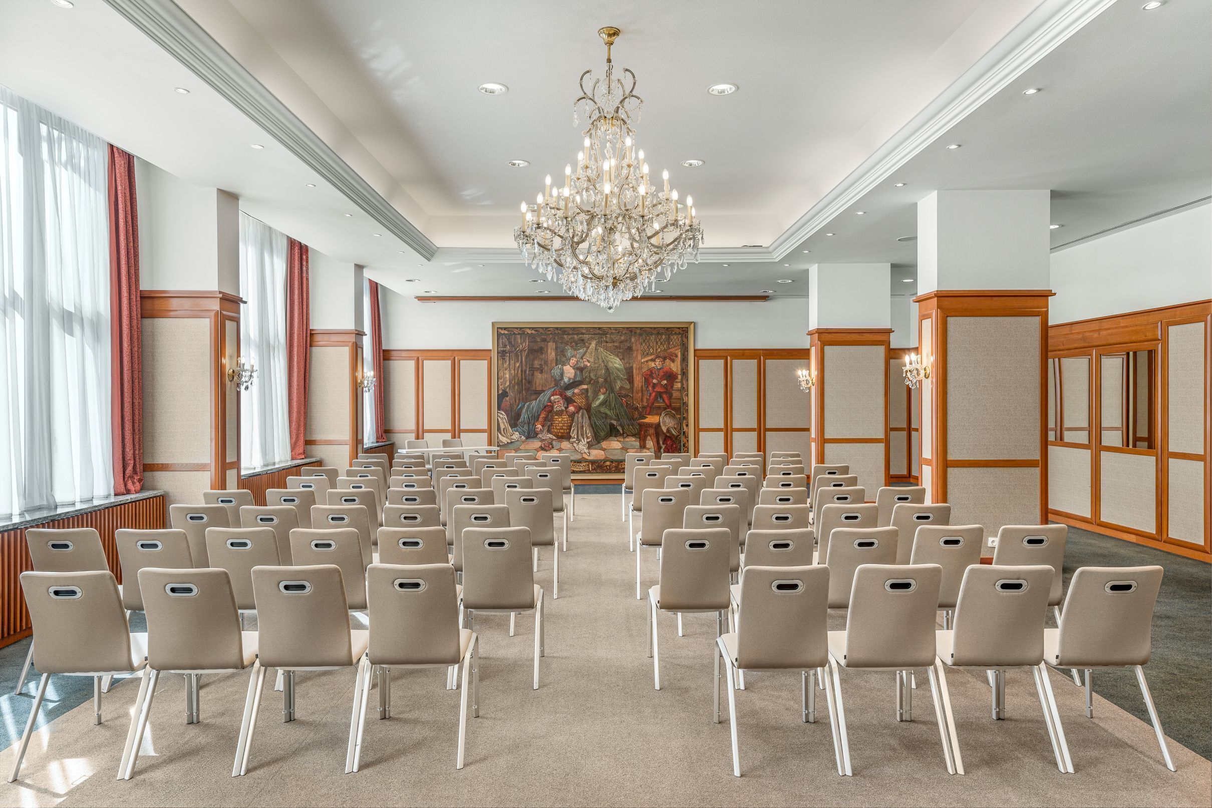 Ein Konferenzraum mit Reihen von Stühlen, einer großen Chandelier und einem Gemälde an der Wand.