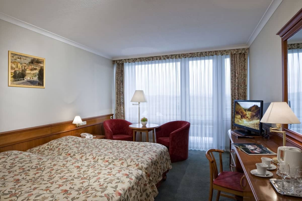 A hotel room with a bed, two red chairs, a desk with a television, and a window with sheer curtains.