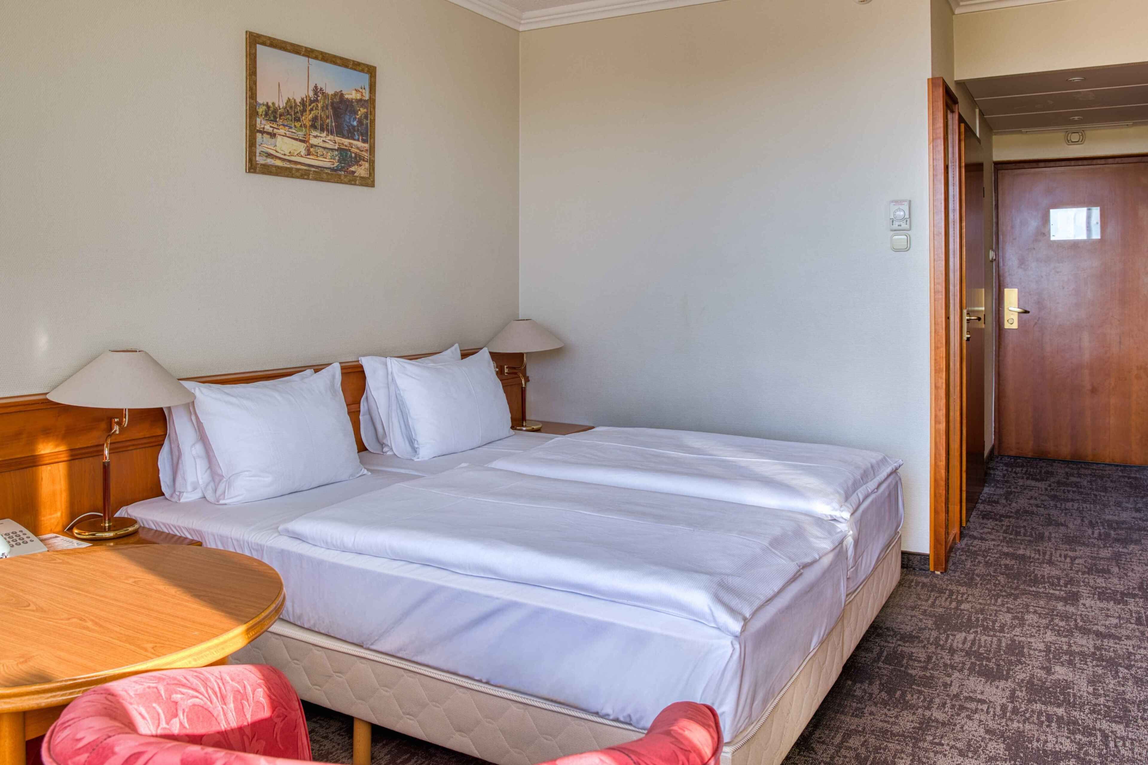 A hotel room with a bed, two bedside tables, a lamp, a painting on the wall, and a door.