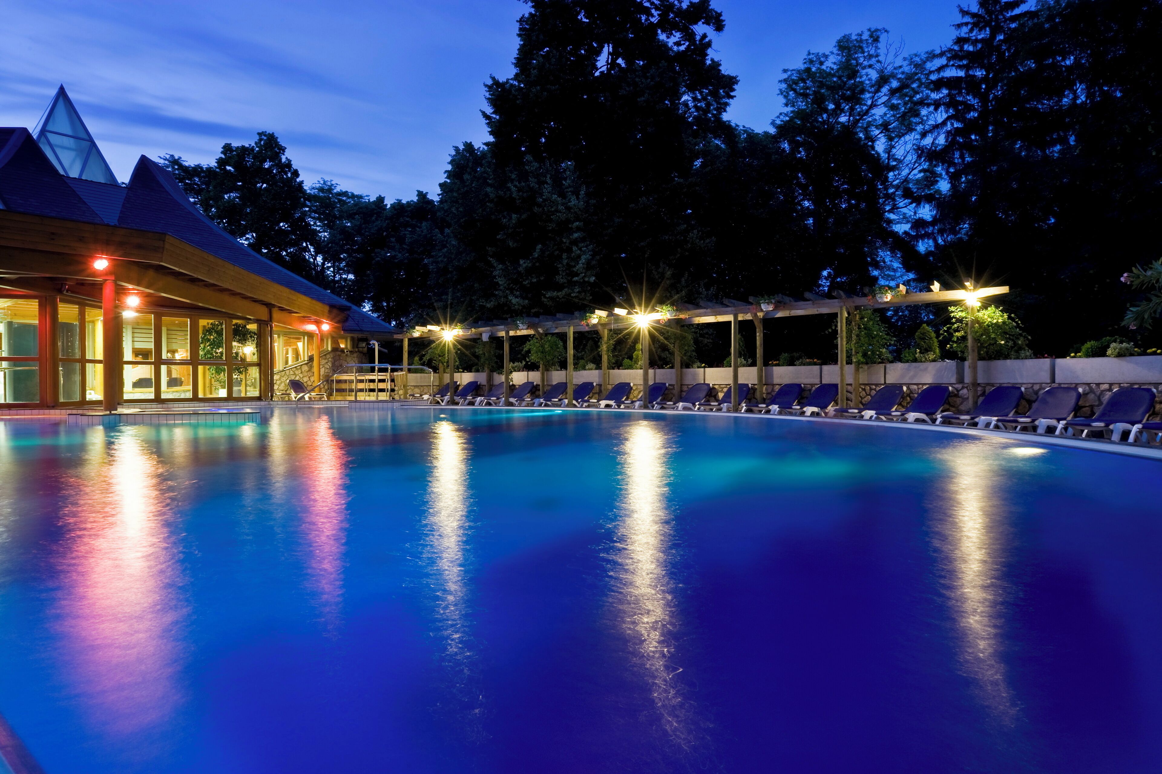 A swimming pool with lounge chairs, a covered area with lights, and a building with a unique roof design.