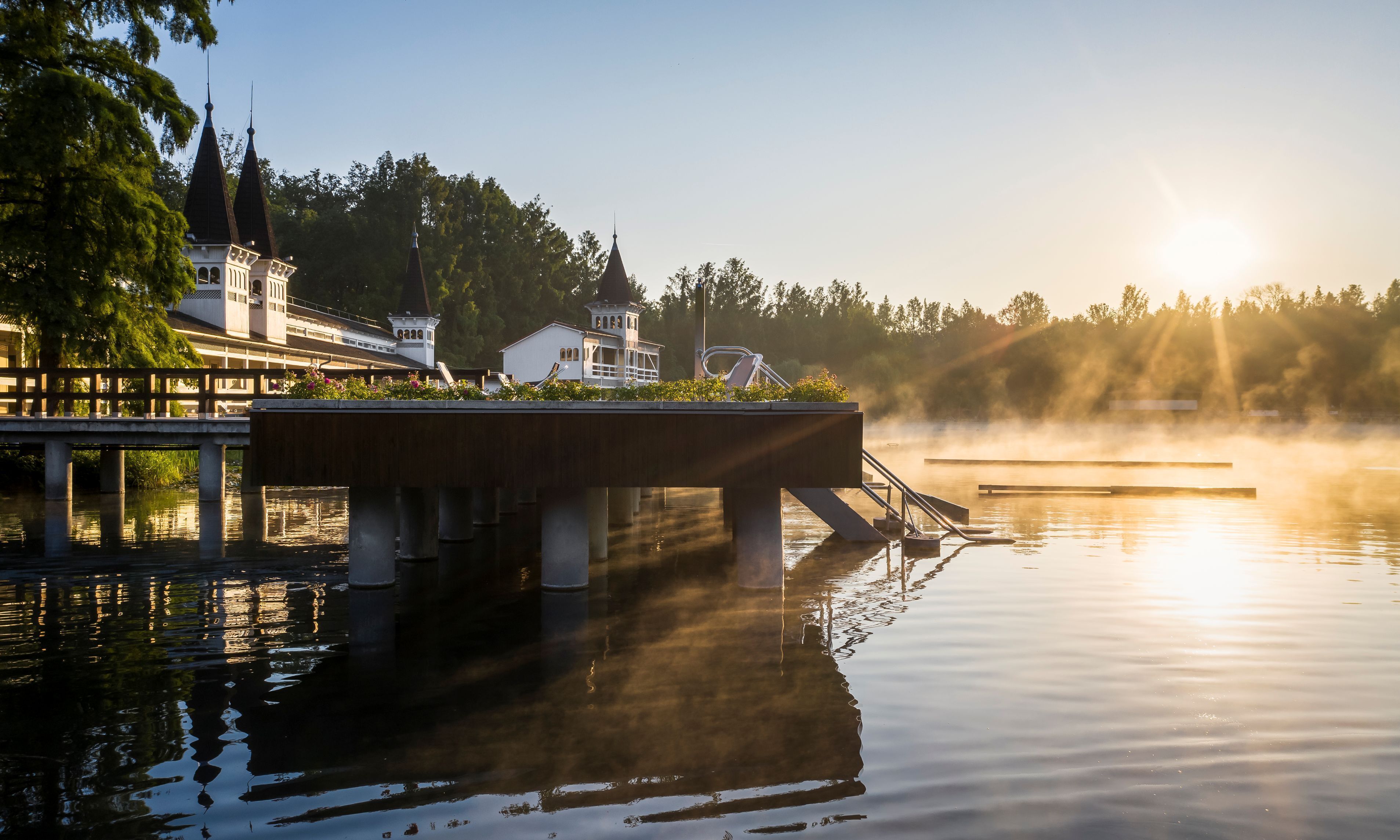 A bridge over calm water with a building on the left and a steeple in the background.
