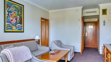 Een hotelkamer met een bank, fauteuil en een houten bijzettafeltje. Aan de muur hangt een schilderij en er is een deur naar een andere kamer.
