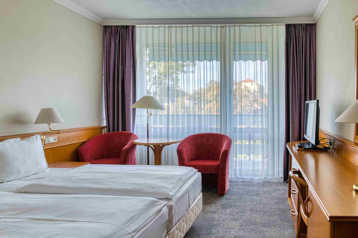 A hotel room with a bed, two red chairs, a small table, a desk with a lamp, and a TV.