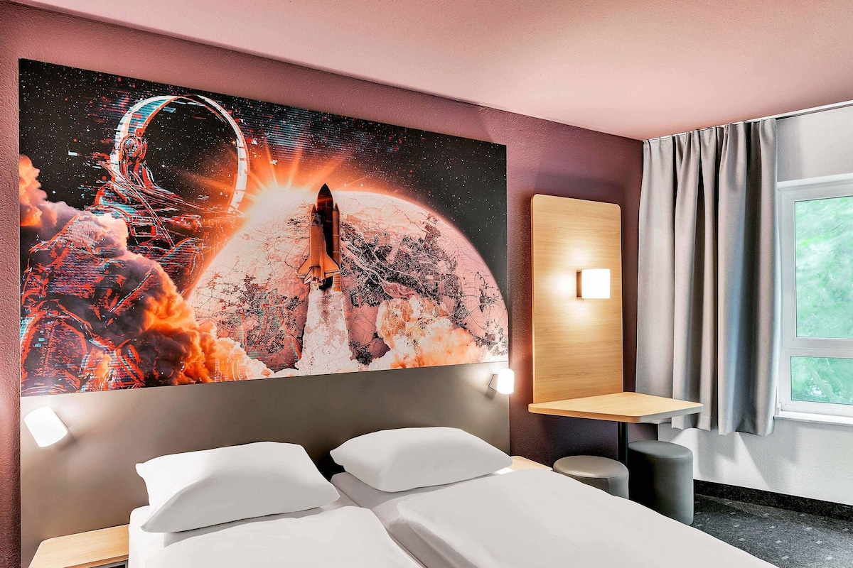 Una habitación de hotel con una amplia obra de arte con temática espacial en la pared, una cama con sábanas blancas, una mesita de noche y un ventanal con cortinas.