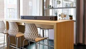 A modern office space with a long wooden desk, three cushioned chairs, and a large window with a view of buildings.