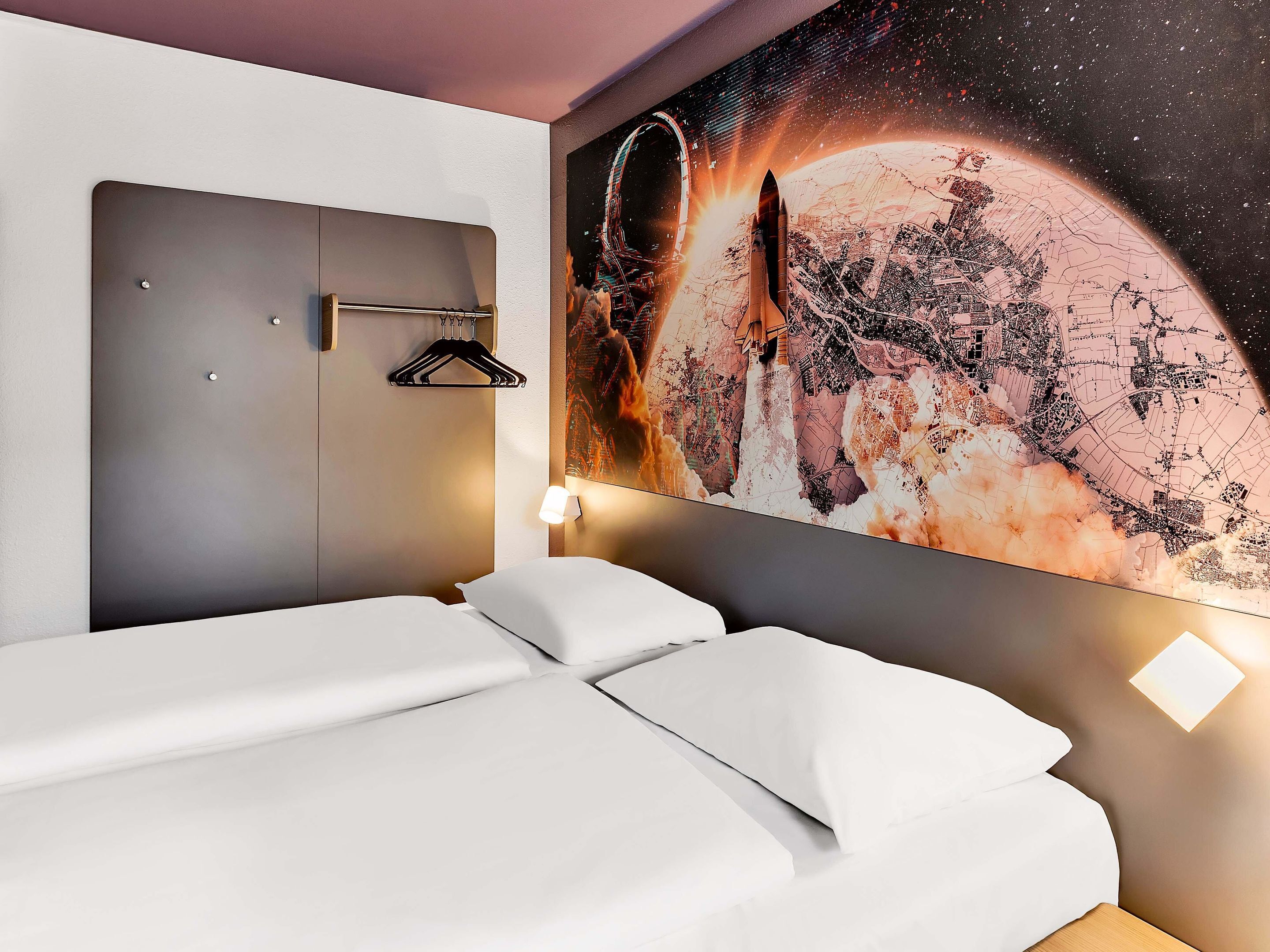 A modern hotel room with a large wall mural of a space scene, a neatly made bed, a wall-mounted hanger, and a bedside lamp.