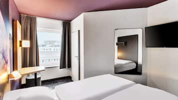 Ein Hotelzimmer mit Bett, Wandfernseher, Fenster mit Vorhängen und Blick auf Gebäude.