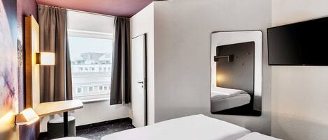 A hotel room with a bed, a TV mounted on the wall, a window with curtains, and a view of buildings outside.