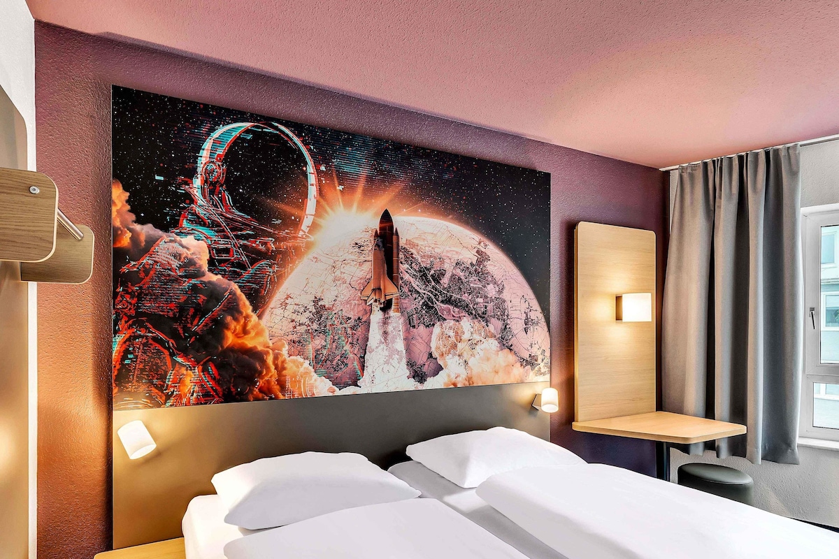 Habitación de hotel con una amplia obra de arte con temática espacial en la pared, cama con ropa blanca, mesita de noche y pequeño escritorio con lámpara.