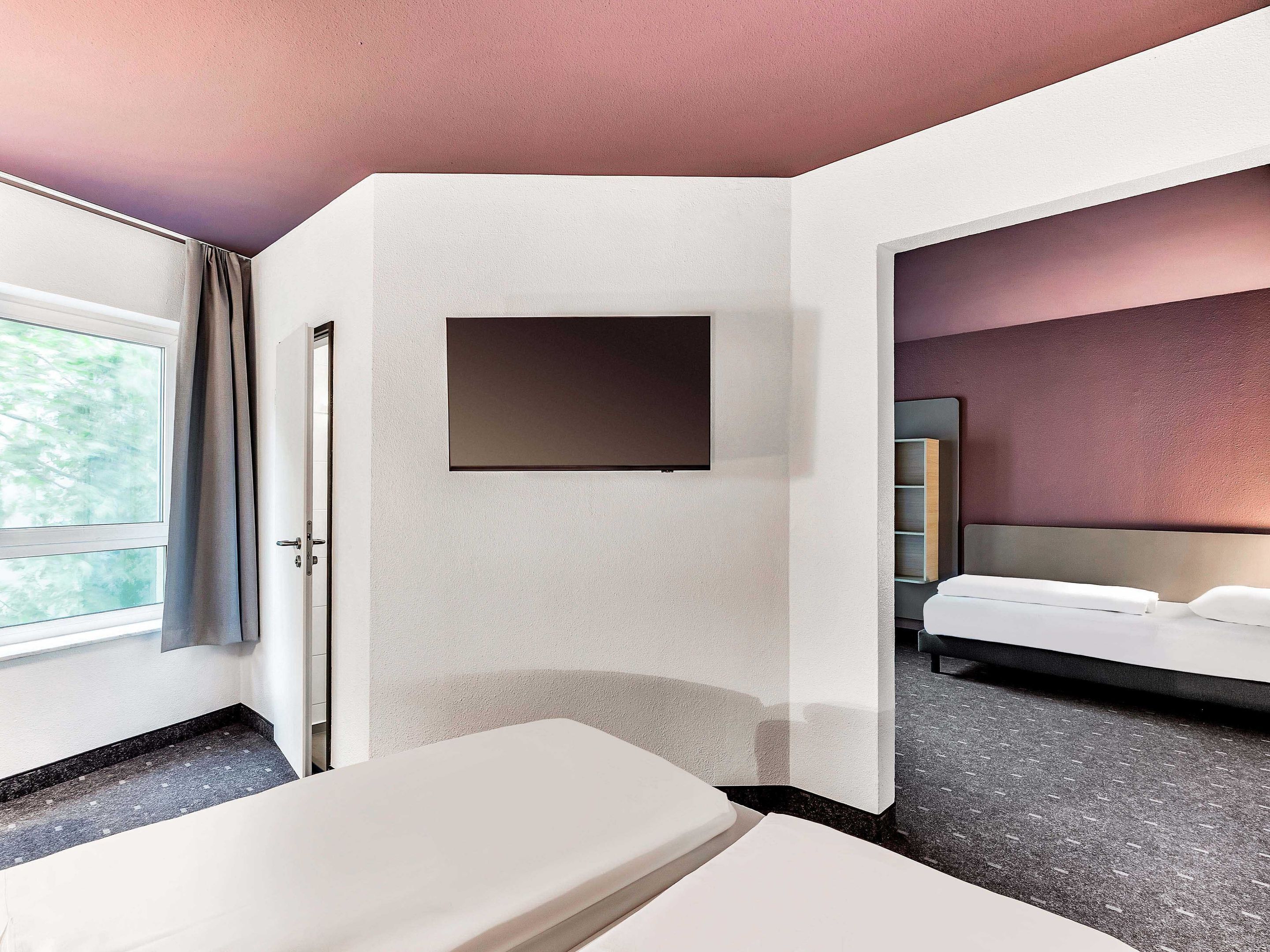 Habitación de hotel con cama, mesita de noche, televisor montado en la pared y ventana con cortinas.