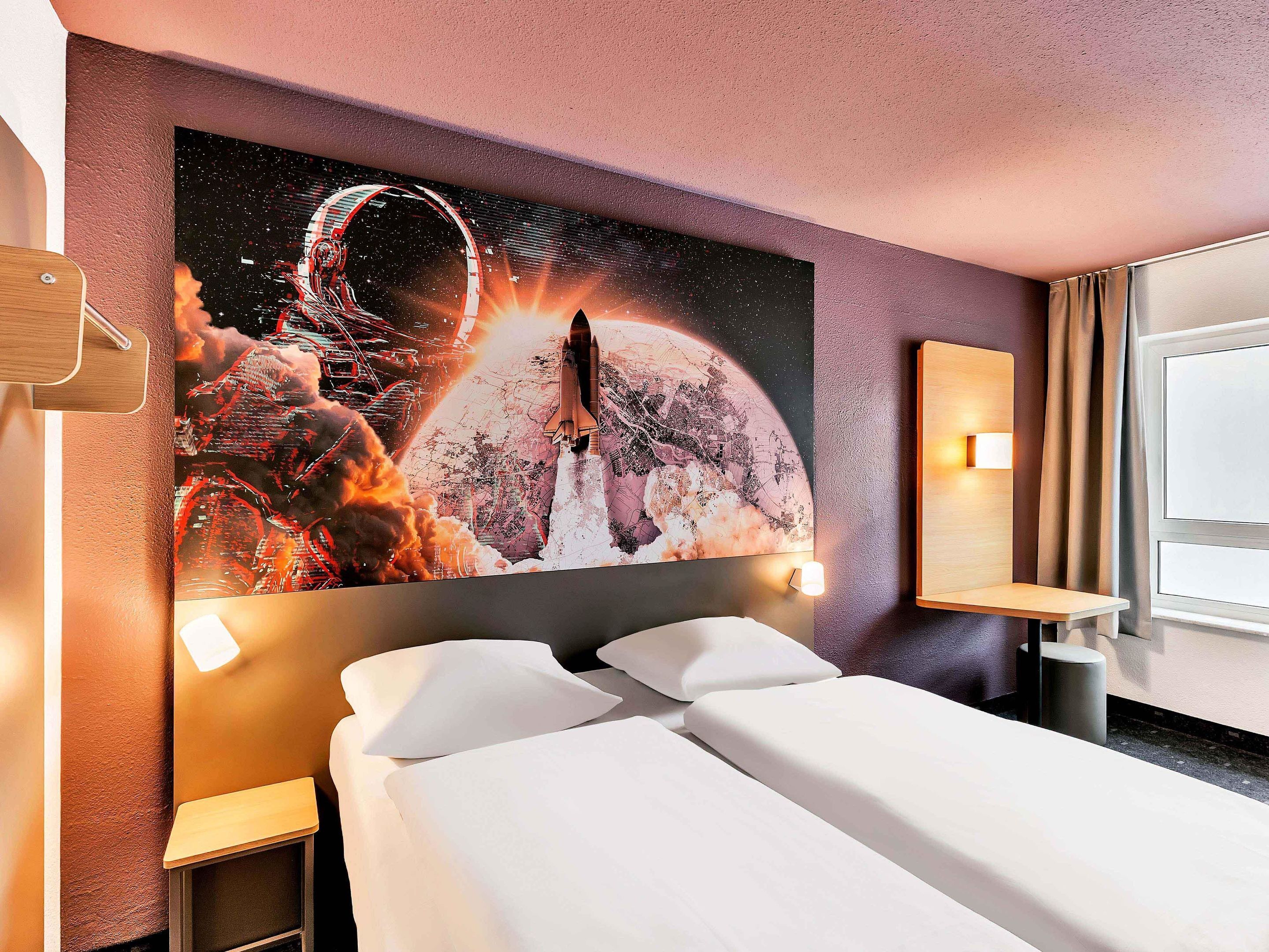A hotel room with a large space-themed mural on the wall, a neatly made bed, and a bedside table with a lamp.