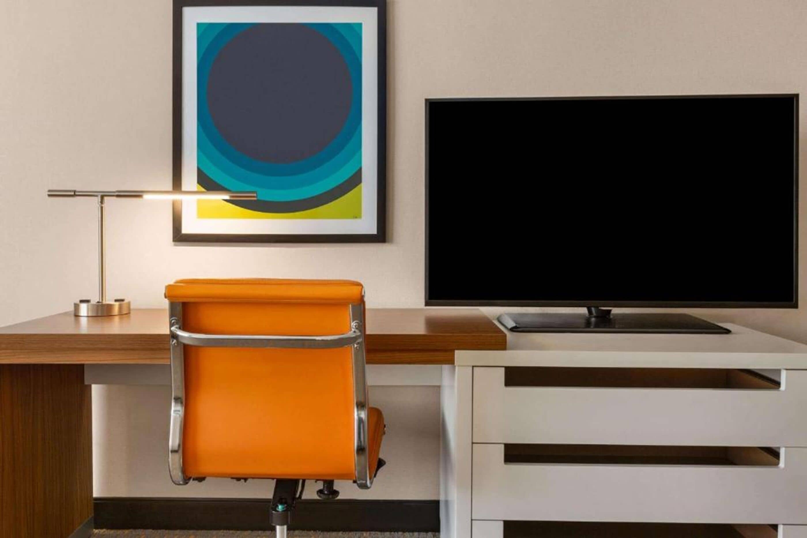 A hotel room with a desk, a chair, a flat-screen TV, and a framed abstract painting.