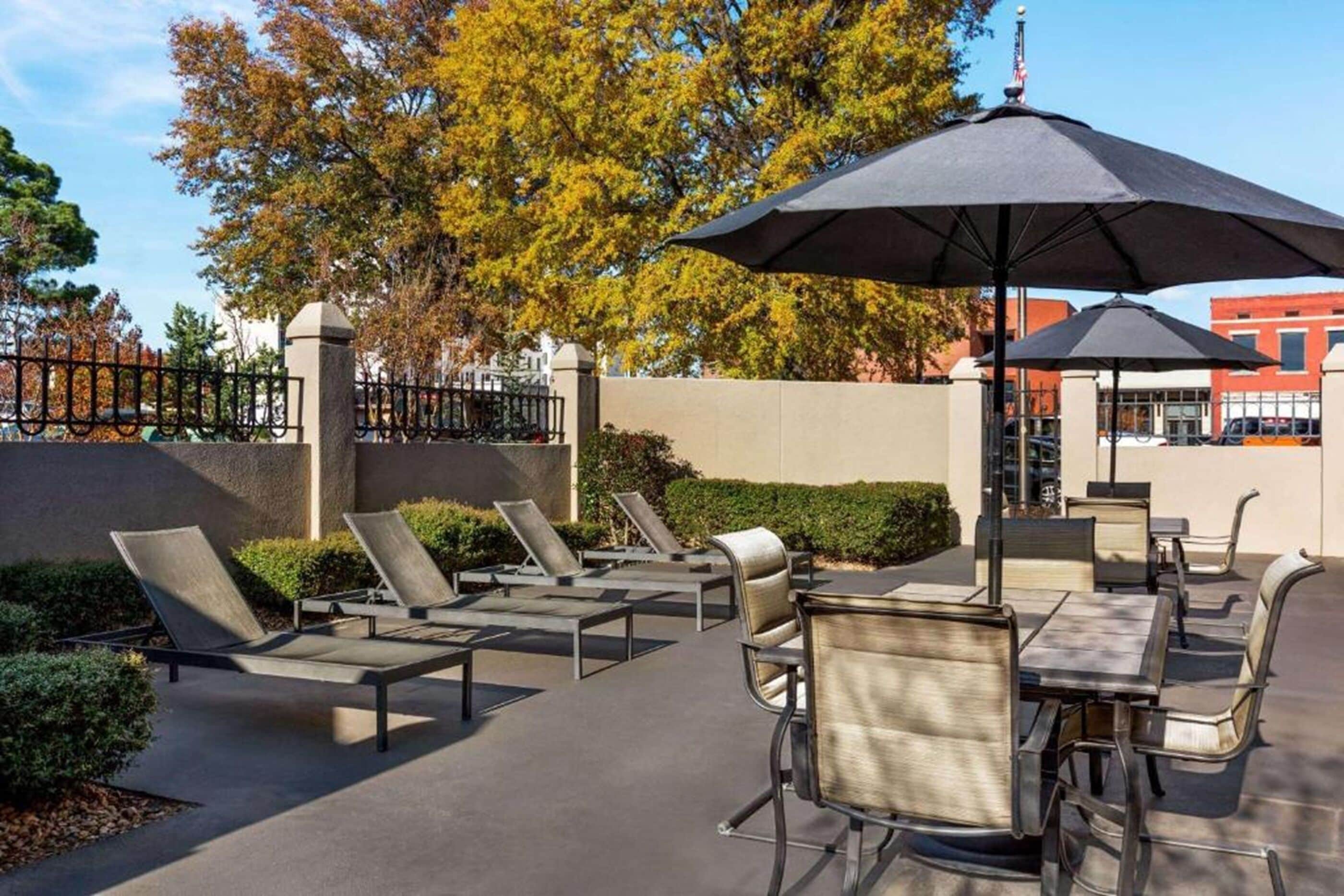 An outdoor patio area with lounge chairs and umbrellas, surrounded by a low wall and trees.
