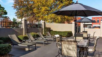 An outdoor patio area with lounge chairs and umbrellas, surrounded by a low wall and trees.
