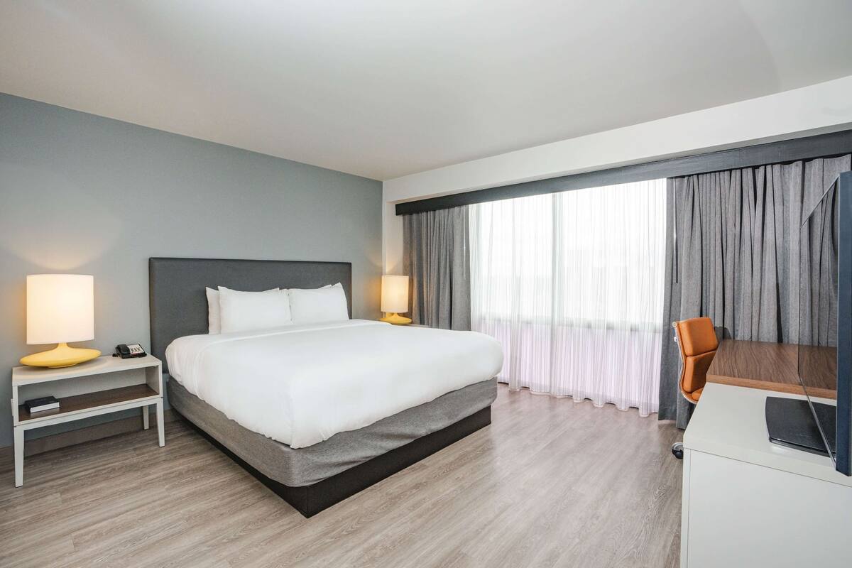 Een hotelkamer met een bed, bedlampen, een bureau met stoel en een groot raam met gordijnen.