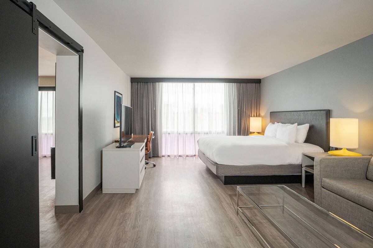 Een hotelkamer met een bed, een bureau, een stoel, een bank en een groot raam met gordijnen.