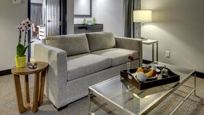 Una habitación de hotel moderna con un sofá gris, una mesa de centro de vidrio con fruta, una mesita auxiliar con una planta y una lámpara.